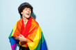 Young happy gay Latin man wear mesh tank top hat clothes wrapped in striped rainbow flag look aside on area isolated on plain blue background studio portrait. Pride day June month love LGBT concept.