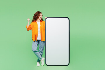 Wall Mural - Full body young woman in orange shirt white t-shirt casual clothes big huge blank screen mobile cell phone smartphone with area do winner gesture isolated on plain green background Lifestyle concept