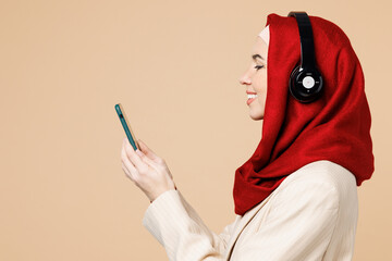 Wall Mural - Side view happy young Arabian Asian Muslim woman wear red abaya hijab suit clothes listen music in headphones use mobile cell phone isolated on plain beige background UAE middle eastern Islam concept