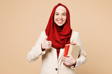 Young smart happy Arabian Asian Muslim woman wear red abaya hijab suit clothes hold in hand books show thumb up isolated on plain beige background studio. UAE middle eastern Islam religious concept.