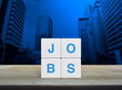 JOBS letter on white block cubes on wooden table over modern office city tower and skyscraper, Business recruitment concept
