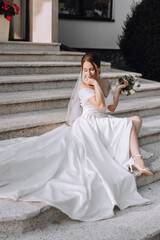 Wall Mural - A woman in a white wedding dress sits on a set of stairs. She is holding a bouquet of flowers in her lap. Concept of elegance and romance, as the bride is dressed in her wedding attire