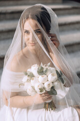 Wall Mural - A bride is sitting on a staircase with a veil covering her face. She is holding a bouquet of flowers in her hands