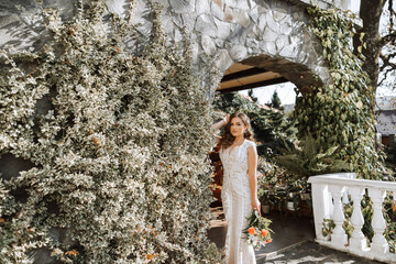 Wall Mural - A woman in a white dress stands in front of a plant with flowers. She is holding a bouquet of flowers in her hand