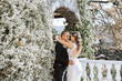 A couple is hugging in front of a white hedge. The man is wearing a black suit and the woman is wearing a white dress. Scene is romantic and intimate