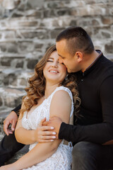 Wall Mural - A man and a woman are sitting on a stone wall, kissing each other. The woman is wearing a white dress and the man is wearing a black shirt. Scene is romantic and intimate