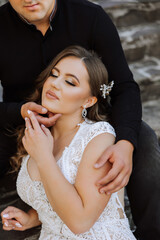 Wall Mural - A woman in a wedding dress is sitting on a chair with her arms around a man. The man is wearing a black shirt and the woman is wearing a white dress. Scene is romantic and intimate