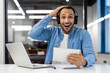 © Tetiana - Portrait of a happy young Muslim man sitting in the office at the table wearing headphones, using a tablet, shocked and surprised holding his hand on his head and smiling at the camera