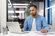 © Tetiana - Serious young hispanic man working in office at desk with laptop and making notes in notebook, watching online training and meeting
