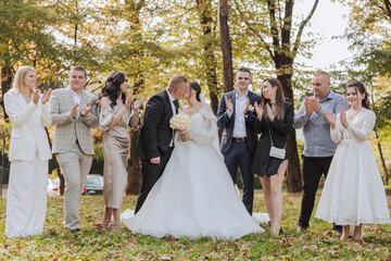 A group of people are posing for a picture, with a bride and groom in the center. The bride is wearing a white dress and the groom is wearing a suit. The group is standing in a park