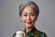 Portrait of an elderly pretty Asian woman with a jar of cosmetic cream in her hand on gray background. Concept of cosmetics for adult people. Silver economy concept.