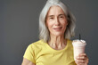 Smiling elderly European woman in yellow t-shirt with glass of protein drink in hand on gray background. Food concept for adult food products, silver economy.