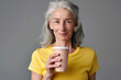 Smiling elderly European woman in yellow t-shirt with glass of protein drink in hand on gray background. Food concept for adult food products, silver economy.