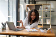 © Wasana - A woman is sitting at a desk with a laptop and pointing at something on the screen. She is smiling and she is happy. The scene suggests a positive and productive atmosphere