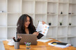 © Wasana - A woman is presenting a report on a laptop in front of a white wall. She is holding a piece of paper and pointing to it. The room has a lot of bookshelves and potted plants