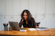 © Wasana - A woman is sitting at a desk with a laptop and a cell phone. She is smiling and she is happy