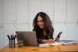 © Wasana - A woman is sitting at a desk with a laptop and a cell phone. She is smiling and she is enjoying her work
