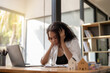 © Wasana - A woman is sitting at a desk with a laptop and a stack of papers. She is looking at the laptop and she is in a state of distress