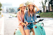 Two young beautiful smiling hipster female in trendy clothes. Carefree women driving retro motorbike on the street background. Positive models having fun, riding classic Italian scooter in Europe city