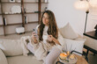 © polinaloves - Young woman using her smartphone and drinking tea, relaxing in a cozy living room.