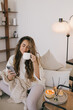 © polinaloves - Young woman using her smartphone and drinking tea, relaxing in a cozy living room.