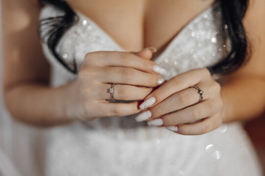 A woman is wearing a white dress and holding her hands up to show off her wedding ring. The ring is a diamond and is set in a gold band. The woman's hands are adorned with nail polish