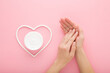 Young adult woman hands and white heart shape with cream jar on light pink table background. Pastel color. Care about clean and soft body skin. Closeup. Point of view shot. Top down view.