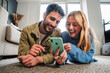 © Jose Calsina - Adult couple watching funny videos together on a cellphone social media app lying on the floor at home. Young caucasian wife and happy husband on the carpet browsing on internet using a smart phone