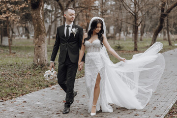 Wall Mural - A bride and groom are walking down a path in a park. The bride is wearing a white dress and the groom is wearing a suit. They are holding hands and the bride is carrying a bouquet