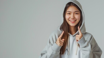 Wall Mural - Smiling Asian young woman in a gray hoodie pointing upwards, looking cheerful on light gray background.