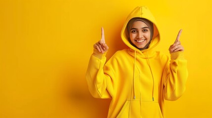 Sticker - Cheerful young woman in yellow hijab and hoodie pointing upwards against a matching yellow background.