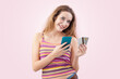 Happy and smiling young woman uses her mobile phone and credit card to browse an internet web page or online store, using her smartphone for shopping and online payments. Isolated on pink background