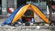 Colorful tent set up on a dirty urban sidewalk surrounded by litter and abandoned items.