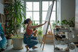 Passionate about process of painting, artist woman applies oil paint with paintbrush to canvas sitting barefoot near window in art studio. Focused painter girl with colour palette drawing on easel.