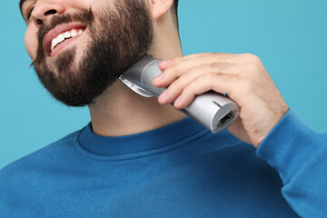 Wall Mural - Handsome young man trimming beard on light blue background, closeup