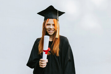 Wall Mural - teenage girl in clothes of a graduate coat and cap celebrates high school or  junior year graduation on background of white wall with shadows and with diploma in hands, education and no school concept