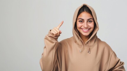 Cheerful young woman in a beige hoodie pointing up, smiling brightly on a light background.