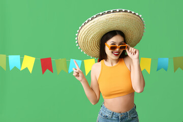 Sticker - Happy young woman in Mexican sombrero hat pointing at garland on green background