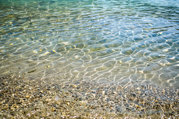 ripples of the crystal clear shallow water surface, on a stony beach of lake outdoor summer backgrou