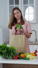 Canvas Print - Young woman unpacking shopping bag, taking out green broccoli. Kitchen background and pretty girl putting fresh vegetables on the table. Healthy food. Vertical video