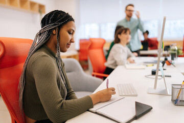 Concentrated African-American woman writing down notes and using computer sitting at work in big modern corporate office. Colleagues businesspeople working together on computer on blurred background.