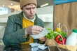 Focused man examining receipts at a cafe table, with fresh groceries including vegetables and milk beside him.