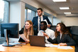 © artiemedvedev - Multiethnic team works in modern office, young woman points at computer screen, presenting ideas. Colleagues gather around desk with laptop, paperwork, engaging in creative group discussion.