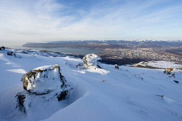 Wall Mural - View from the mountain to the city of Magadan. In the foreground is a snow-covered mountain slope and snow-covered rocks. In the distance is a large northern city. Magadan region, Far East of Russia.
