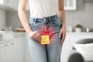 Woman holding sticky note with drawn sad face and suffering from cystitis symptoms indoors, closeup