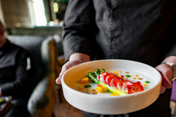 Wall Mural - A person in a dark shirt holds a plate of delicious gourmet dish featuring asparagus and colorful garnish in a cozy interior setting