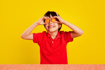 Cheerful little boy in red polo shirt using carrot slices as pretend glasses against yellow background. Creative fun with food. Concept of food, childhood, emotions, meal, menu, pop art