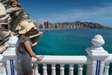 Fototapeta  - Woman standing on the balcony (Balcó del Mediterrani) in a hat and khaki dress (wind in the hair, holiday atmosphere) looking towards the Spanish Manhattan (Benidorm)