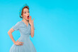shocked woman, pin-up girl with hairstyle covering opened mouth with hand on blue background with copy space