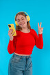 Cheerful curvy woman carried away with music dances carefree with phone in hands with  headphones on ears on blue background